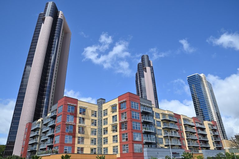 Edificios residenciales modernos de colores rojo y crema frente a rascacielos de cristal bajo un cielo azul despejado.